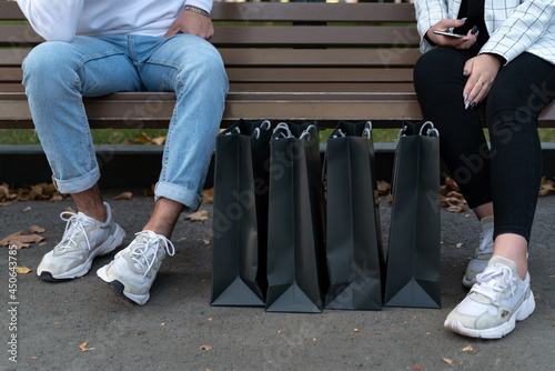 Paper shopping bags next to shoppers resting on bench. Feet of young man and woman after shopping