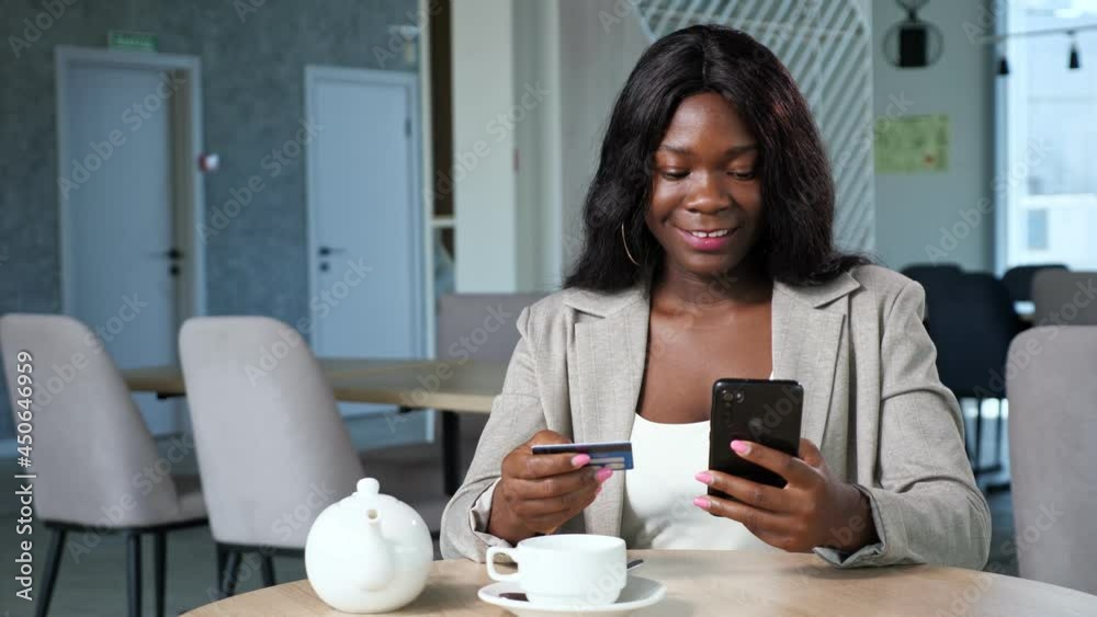 Cheerful black woman in stylish jacket pays online with credit card and mobile phone sitting at table with teapot and cup in cozy cafeteria Cheerful black woman in stylish jacket pays online with credit card and mobile phone sitting at table with teapot and cup in cozy cafeteria