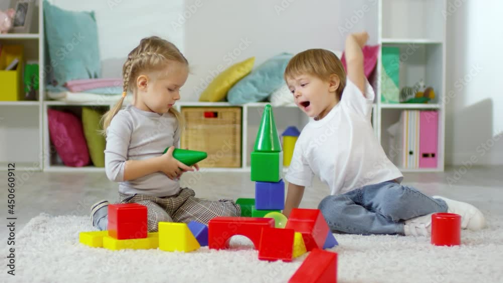 Cute kids playing educational game on carpet in home: little girl building toy tower with colorful plastic blocks while her playful brother destroying it Cute kids playing educational game on carpet in home: little girl building toy tower with colorful plastic blocks while her playful brother destroying it