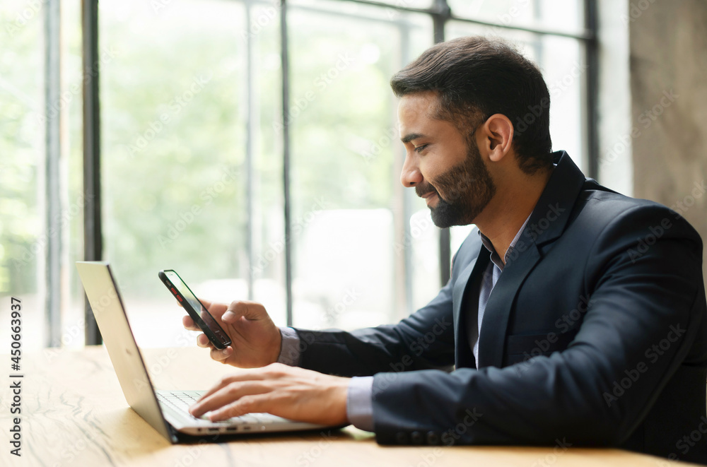 Smiling Indian male office employee in smart casual wear using smartphone during online work on the laptop, chatting online in social medias sitting at the desk in the office, side view Smiling Indian male office employee in smart casual wear using smartphone during online work on the laptop, chatting online in social medias sitting at the desk in the office, side view