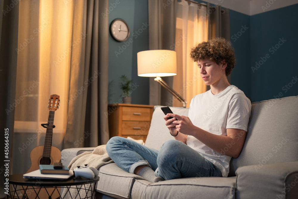 A bored young boy, teenager, schoolboy, student is relaxing on the couch in the evening with his phone, smartphone in hand. The young boy sends messages to friends, browses social media A bored young boy, teenager, schoolboy, student is relaxing on the couch in the evening with his phone, smartphone in hand. The young boy sends messages to friends, browses social media