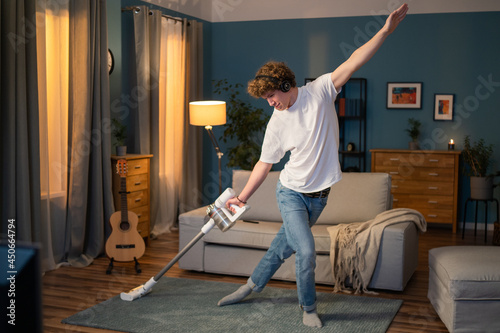 A young boy is cleaning the living room in the evening. The teenager has fun vacuuming the carpet in the room, listening to music on wireless headphones and dancing with the vacuum cleaner.