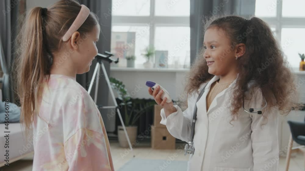 Handheld tracking shot of giggling 8-year-old girls playing doctor at home. One of them is wearing white coat and stethoscope and measuring temperature of her cheerful friend Handheld tracking shot of giggling 8-year-old girls playing doctor at home. One of them is wearing white coat and stethoscope and measuring temperature of her cheerful friend