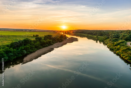 Scenic view at beautiful summer river sunset with reflection on water with green bushes, grass, golden sun rays, calm water ,deep blue cloudy sky and glow on a background, spring evening landscape © Yaroslav