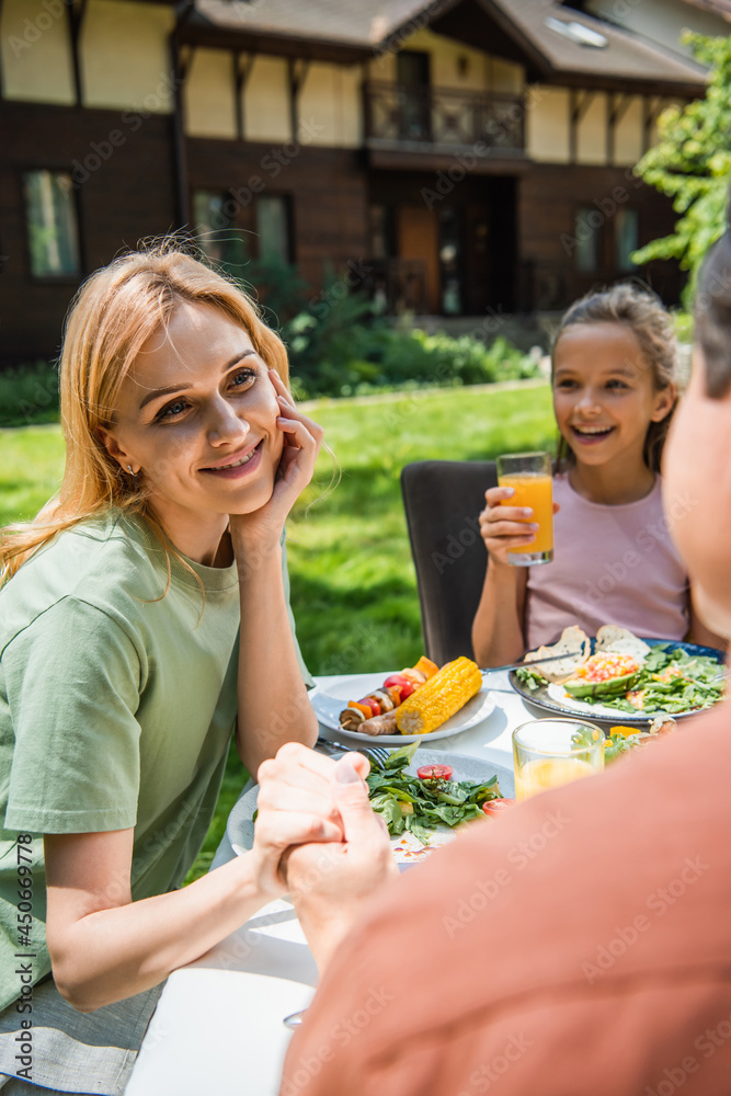 Smiling woman holding hand of blurred husband near cheerful kid with orange juice and food outdoors Smiling woman holding hand of blurred husband near cheerful kid with orange juice and food outdoors