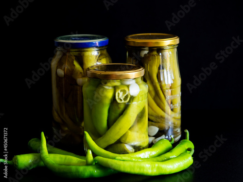 In this photoillustration, homemade pickled chili peppers in glass jars on black background