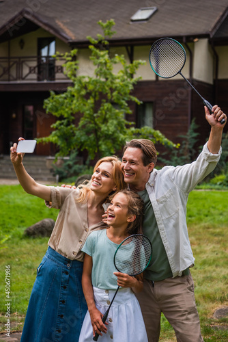 Smiling woman taking selfie on smartphone near cheerful family with badminton rackets