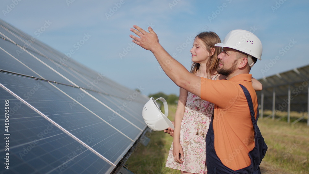 A father, a solar power engineer, and his daughter are standing near solar panels. The father explains to the child the principle of solar electricity and puts a protective helmet on the girl's head. A father, a solar power engineer, and his daughter are standing near solar panels. The father explains to the child the principle of solar electricity and puts a protective helmet on the girl's head.