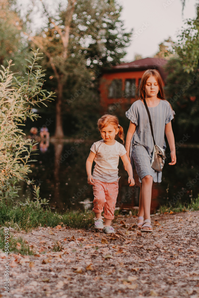 Fototapeta premium Two little girls enjoy summer in park with pond. Big sister holding hand of younger one. Summer countryside walk.