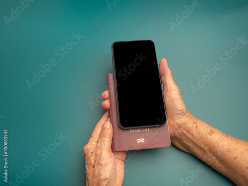 Hands of a senior woman holding a passport and smartphone on a green background. Space for text. Close-up photo. Aged people, communication and travel concept