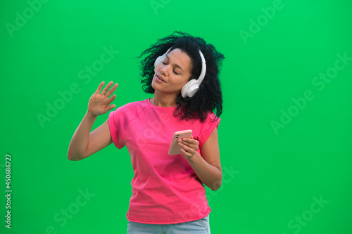 Portrait of young female African American enjoying music in big white headphones using smartphone. Black woman with curly hair poses on green screen in the studio. Close up.
