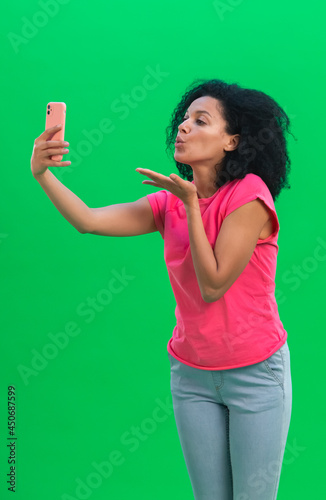 Portrait of young female African American talking for video chat using mobile phone and blows a kiss. Black woman with curly hair in pink tshirt and jeans poses on green screen in the studio. Close up
