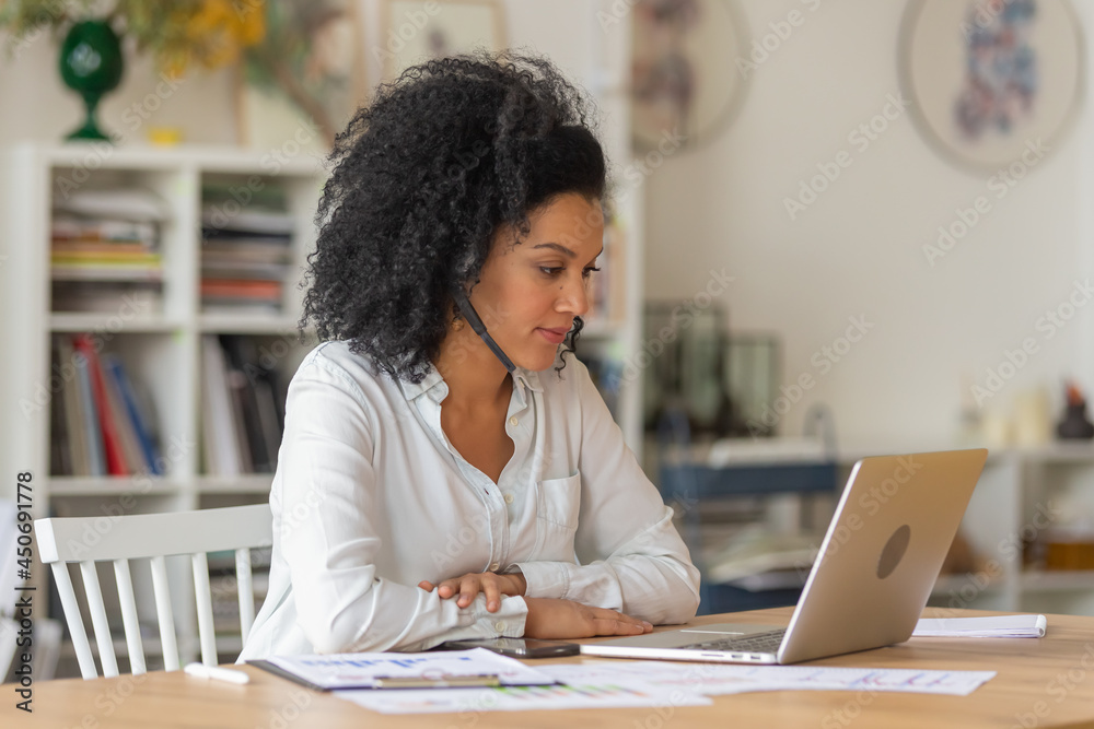 Portrait of a young African American woman talking on video conference call using laptop and headset. Brunette with curly hair in white blouse sits at table in light home office. Close up. Portrait of a young African American woman talking on video conference call using laptop and headset. Brunette with curly hair in white blouse sits at table in light home office. Close up.