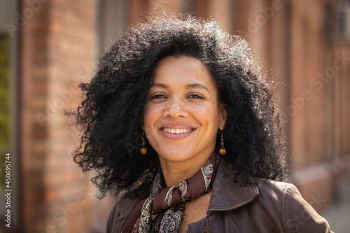 Portrait of stylish young African American woman smiling and looking at camera. Brunette with curly hair in brown leather jacket posing on street against backdrop of blurred brick building. Close up.