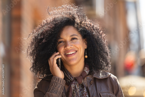 Portrait of young African American woman talking on her smartphone and smiling. Brunette with curly hair in leather jacket posing on street against backdrop of beautiful brick building. Close up.