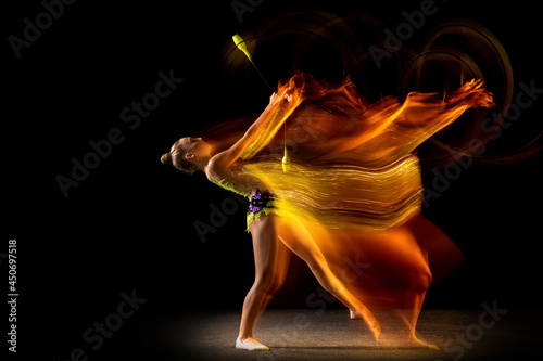 Portrait of young girl, rhythmic gymnastics artist in action isolated on dark studio background with mixed light. Concept of sport, action, aspiration, beauty