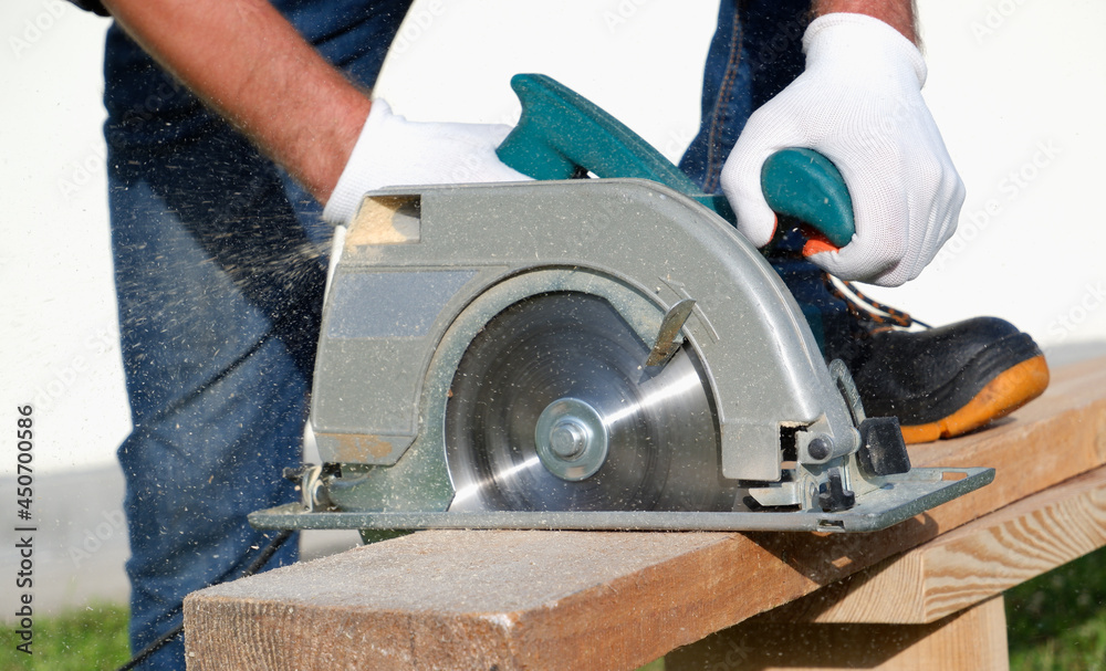 A male joiner works with a hand-held circular saw. A builder is sawing a board at the construction site of a new house. A male joiner works with a hand-held circular saw. A builder is sawing a board at the construction site of a new house.