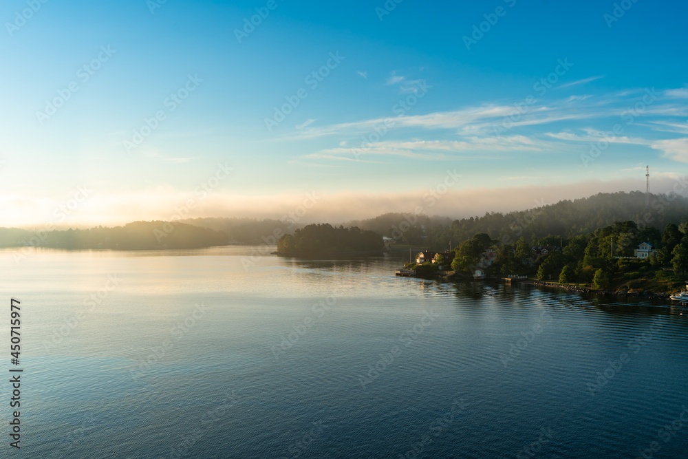 Golden light of the rising sun in the morning over forest islands of archipelago. Magic misty morning. Beautiful mystical clouds enveloped the shores of Scandinavia. View of coast from cruise ship.
