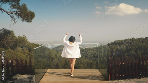 Woman on viewpoint looks on mountain forest landscape, back view. Young female in white goes to cliff observation deck. Summer vacation. Travel destinations, outdoor tourism. Drone flight slow motion photo