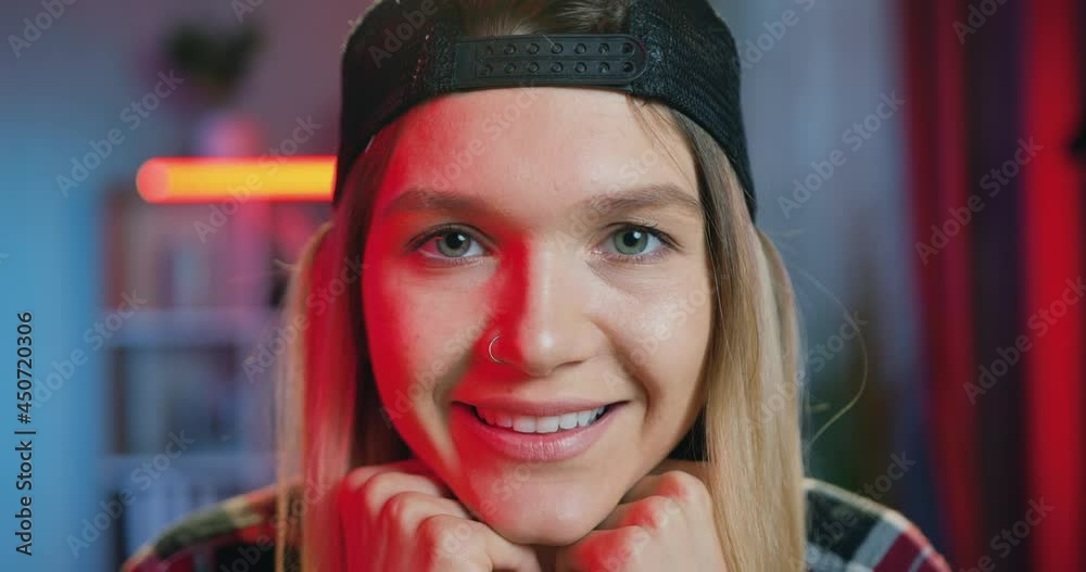Close up of attractive happy relaxed modern young blondie in cap with earring in nose which looking into camera on the background of night lighted room Close up of attractive happy relaxed modern young blondie in cap with earring in nose which looking into camera on the background of night lighted room