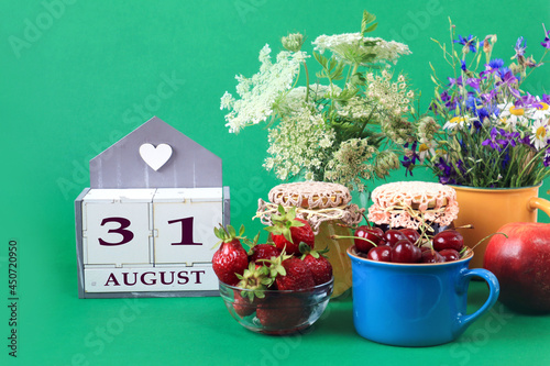 Calendar for August 31 : the name of the month of August in English, cubes with the number 31, bouquets of wild flowers, jars of jam, strawberries and cherries in cups, green background © MARYIA