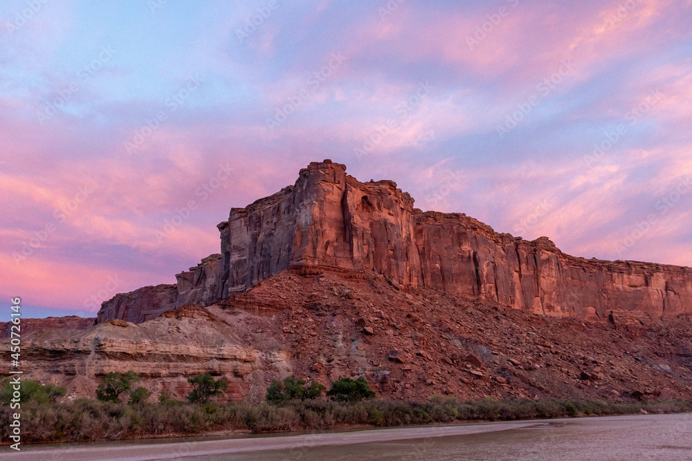 The brilliant colors of a sunrise reflecting on clouds over the top of a canyon wall with a river in the foreground in Utah.