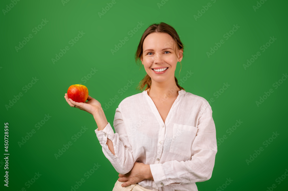 A middle-aged woman, forty years old, smiles and looks at the camera holding a fresh apple in her hand in a white shirt on a green background. A middle-aged woman, forty years old, smiles and looks at the camera holding a fresh apple in her hand in a white shirt on a green background.