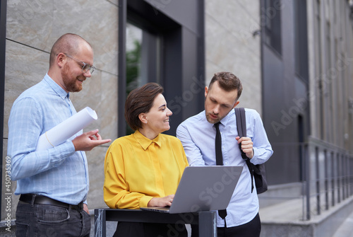 Designers or architect team looking at project blueprint and laptop as presentation outdoors. Business persons, men and woman in casual clothes standing outside, brainstorming project