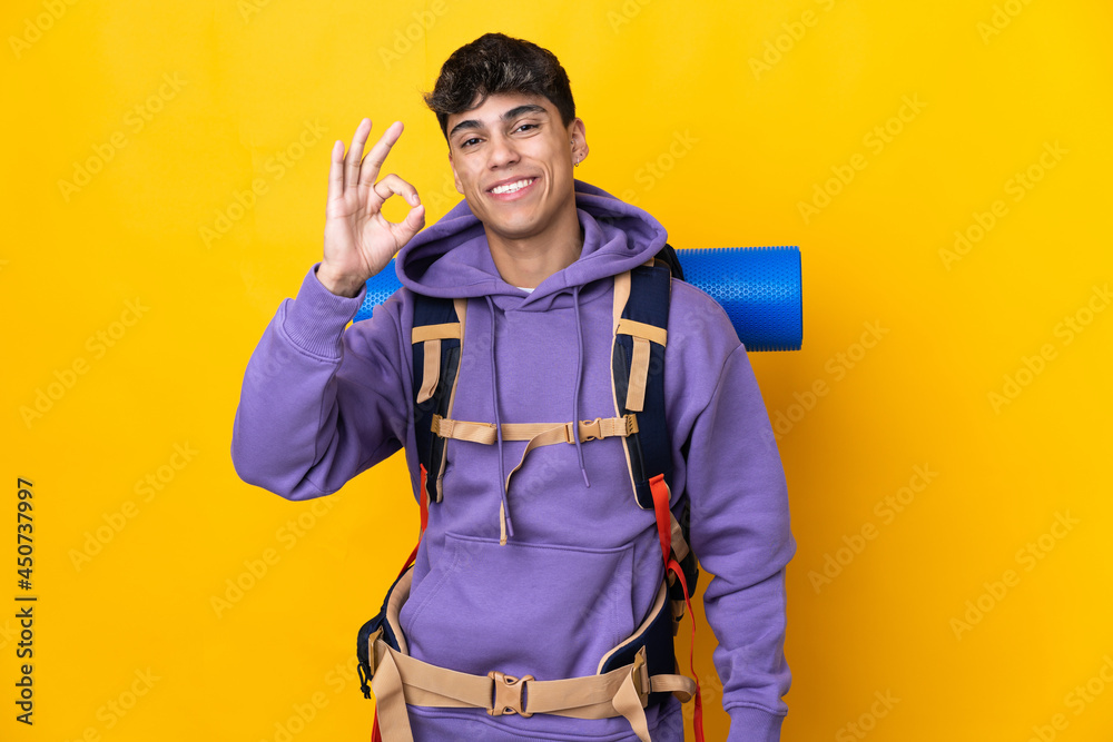 Young mountaineer man with a big backpack over isolated yellow background showing ok sign with fingers