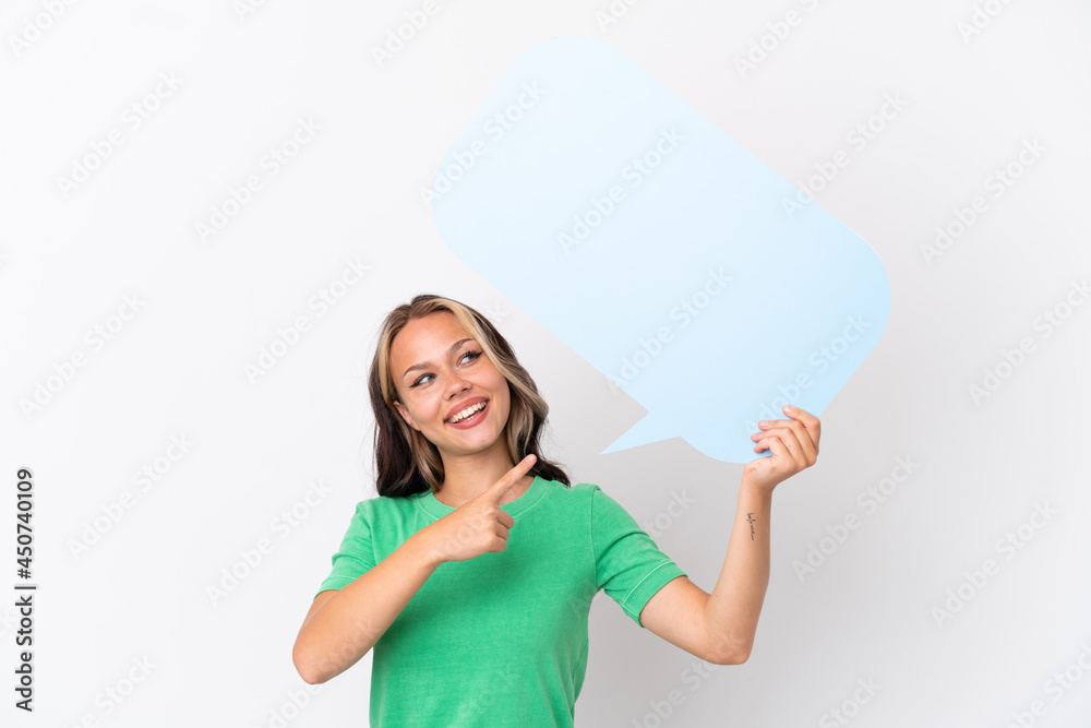 Teenager Russian girl isolated on blue background holding an empty speech bubble and pointing it Teenager Russian girl isolated on blue background holding an empty speech bubble and pointing it