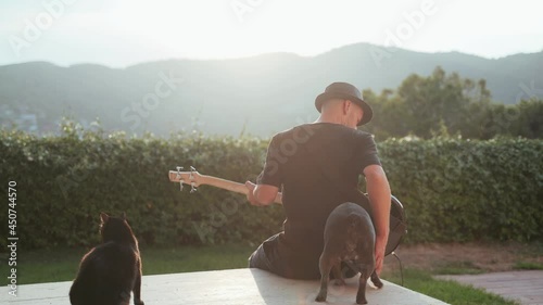 Man in a black hat plays the bass guitar. Musician and his black cat and grey french bulldog on the terrace with a beautiful view. Bass player enjoying the golden evening with his pets. Funny dog 