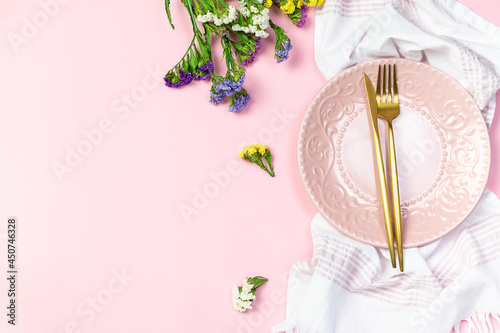 Festive table setting with pink plate, golden cutlery, flowers and napkin on light pink background with copy space for your design.