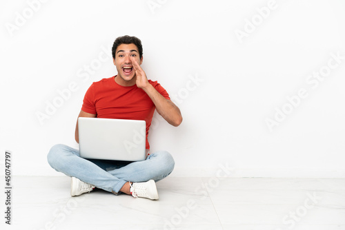 Caucasian handsome man with a laptop sitting on the floor shouting with mouth wide open