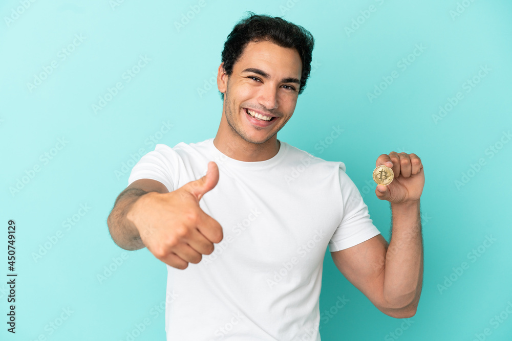 Young man holding a Bitcoin over isolated blue background with thumbs up because something good has happened Young man holding a Bitcoin over isolated blue background with thumbs up because something good has happened