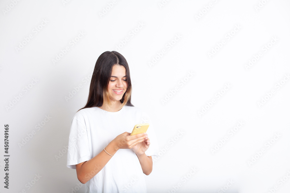 A beautiful teenage girl looks at the smartphone screen and smiles. Joy, happiness. Social networks, content. Communication. SMS. Good news. Like. Place for text, white background. A beautiful teenage girl looks at the smartphone screen and smiles. Joy, happiness. Social networks, content. Communication. SMS. Good news. Like. Place for text, white background.