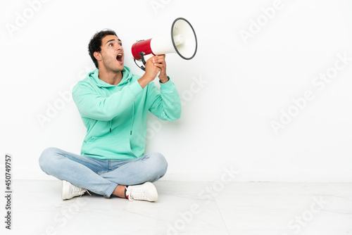 Caucasian handsome man sitting on the floor shouting through a megaphone