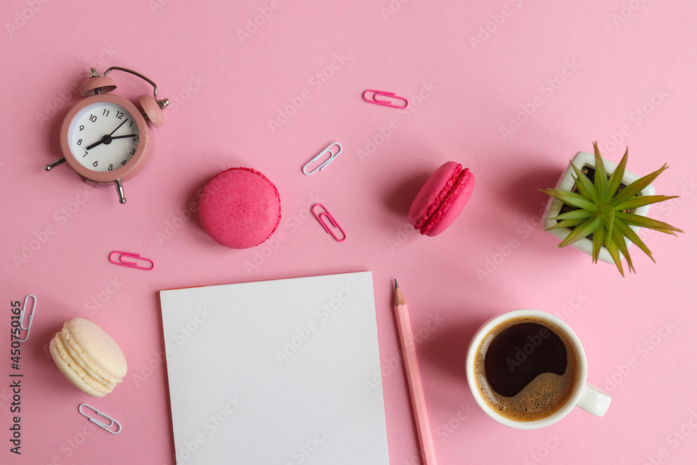 Workplace with notepad, pencil, alarm clock, plant, a cup of coffee and macarons on a pink background. Business, freelance and education concept. Morning time. Top view, flat lay