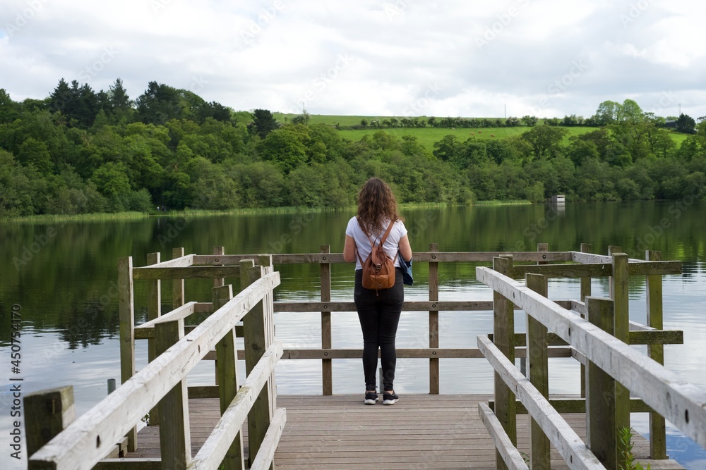 Young traveler with a backpack in lake dock enjoying the landscape. Young woman tourist watching green mountain in Ireland