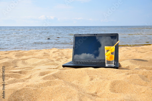 Remote work, a black laptop and a glass of fresh juice on a sandy beach.