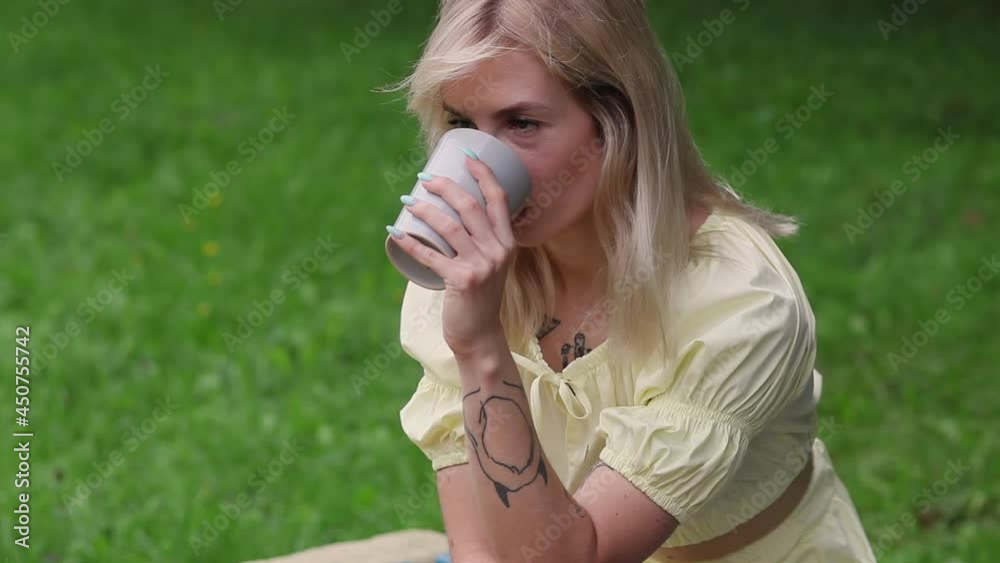 A beautiful girl on a bright summer day drinks tea or coffee while sitting on a park bench. Stylish hipster girl sits on a bench outdoors. She is enjoying a drink. A beautiful girl on a bright summer day drinks tea or coffee while sitting on a park bench. Stylish hipster girl sits on a bench outdoors. She is enjoying a drink.
