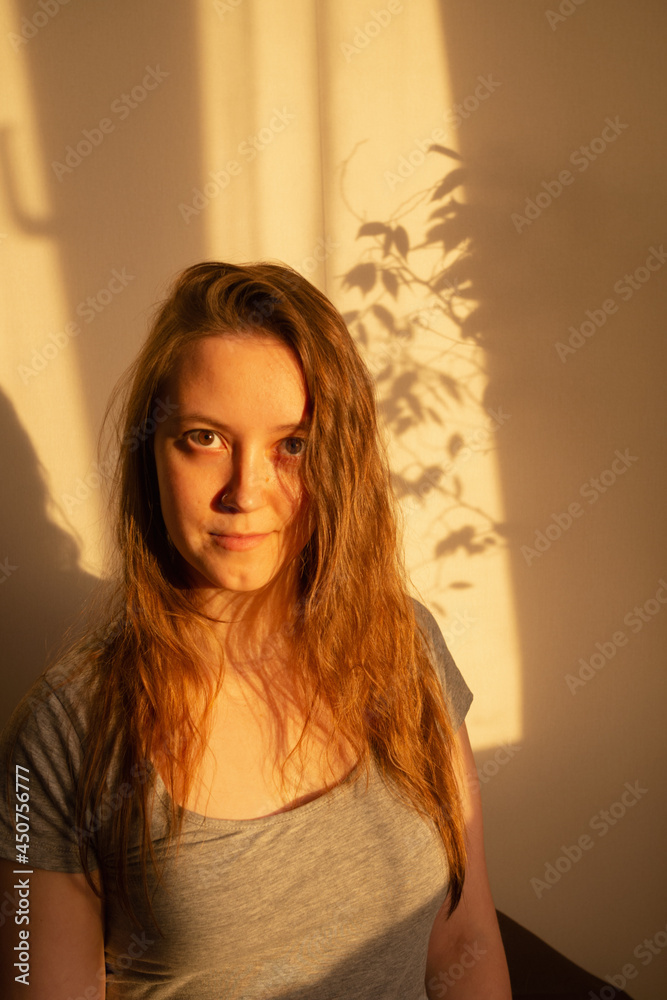 Young woman looking at camera in warm rays of sunset. Warm calm portrait of girl indoor. light and shadow on the face Young woman looking at camera in warm rays of sunset. Warm calm portrait of girl indoor. light and shadow on the face