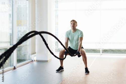 Caucasian blonde man training with rope in functional training fitness gym with big windows and a lot of daylight. photo