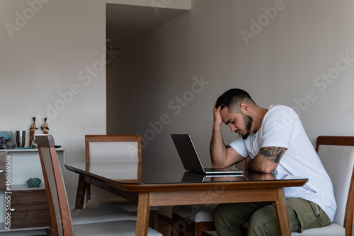 young latin tattooed man in front of laptop stressed out and holding his head