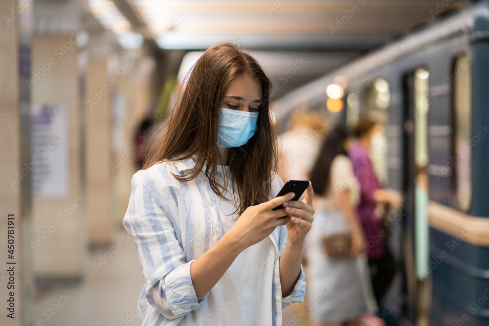Young caucasian woman wear protective face mask, using mobile smart phone, chatting in social media, waiting for train on subway station platform during pandemic Covid-19. New normal concept. 
