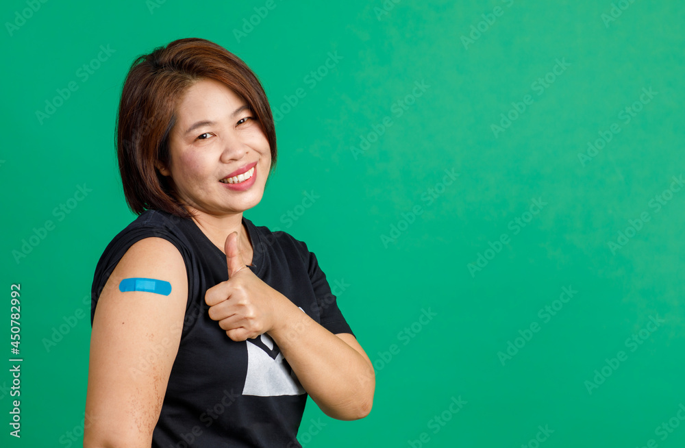 Fototapeta premium Studio shot of Asian middle aged female patient sit smile look at camera show thumb up and blue plaster bandage on arm after receive coronavirus covid 19 vaccination at clinic on green background