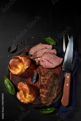 Roastbeef, yorkshire puddnings, basil leaves on a wooden cutting bords with napkin, fork and knife. Rustic style.