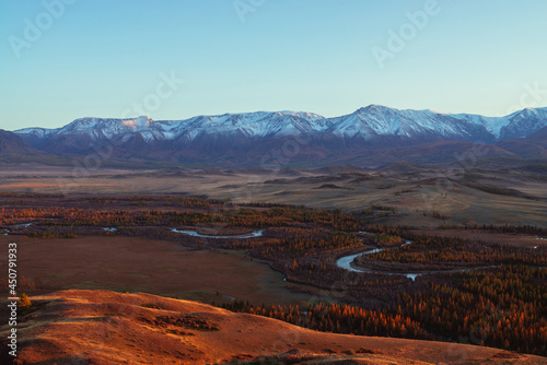 Awesome autumn landscape with mountain river in forest valley and great snow-covered mountain range in red sunset sunshine. Spectacular view from hill to snowy mountains and autumn valley in sunlight.