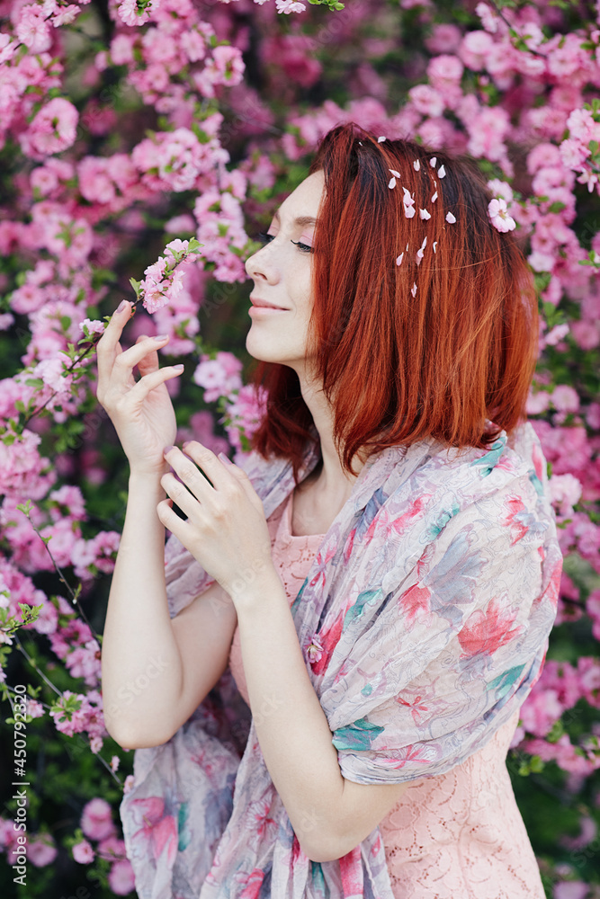 Naklejka premium Russian woman fashion close-up portrait. Beautiful sensual Caucasian young girl perfect skin creartive artistic makeup fashionable spring dress, short red hair. Girl standing in front of a sakura tree