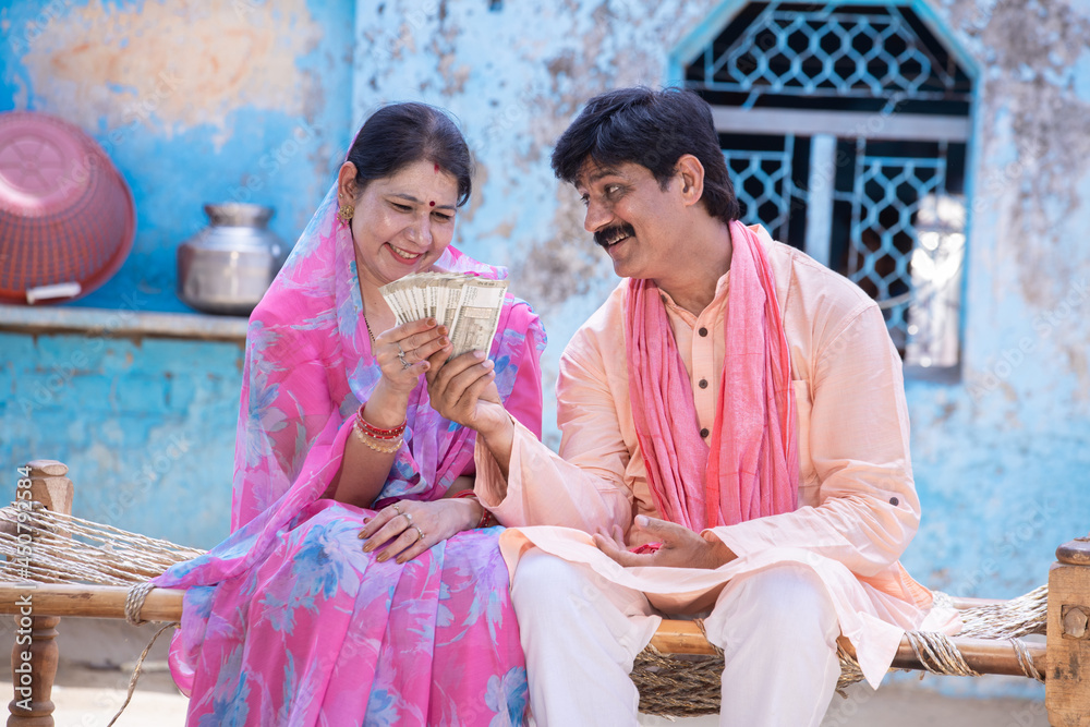 Happy rural woman in sari holding counting indian rupees note, farmer man give cash money to woman, husband and wife sit on bed outside village house. savings and financial growth.