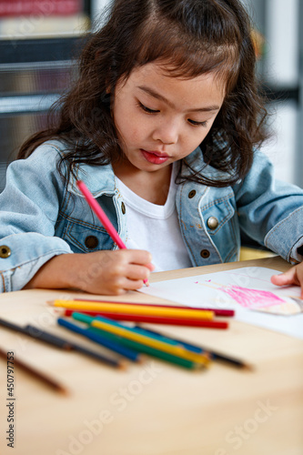 Little Asian girl concentrate to create picture drawing for school homework by pink pencil as art education while sitting on working desk of colored painting tools in reading room at home